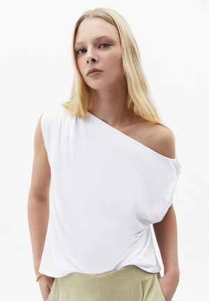 Jeune femme aux longs cheveux blonds portant un top blanc asymétrique et un pantalon beige, debout devant un fond clair uni.