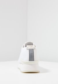 Chaussure de sport blanche avec une tige en mesh texturé, une semelle intermédiaire crème, et une languette arrière ornée d'un logo et d'accents à rayures bleu marine.