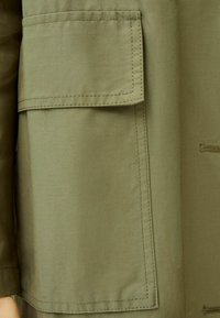 Tela verde oliva con una textura suave, con un bolsillo tipo parche y detalles de costura visibles, que ofrecen un diseño funcional y estructurado.