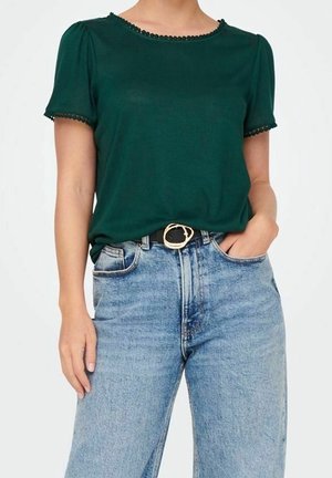 Frau trägt ein dunkelgrünes Kurzarmoberteil mit Spitzenbesatz und hellblaue hoch taillierte Jeans mit schwarzem Gürtel und goldener Schnalle.