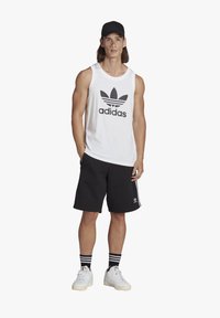 Balta marškinėliai be rankovių su juodu Adidas logotipu, juodos šortai, balti sportbačiai ir juoda kepurė. Ypatybės - juodai baltos dryžuotos kojinės.