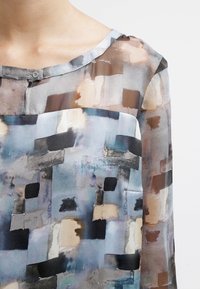 Blusa trasparente con scollatura rotonda, caratterizzata da un motivo astratto multicolore in toni delicati di blu, marrone e beige. Tessuto morbido.