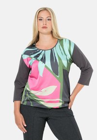 Vaaleatukkainen nainen, joka pukeutunut moniväriseen pitkähihaiseen yläosaan, jossa on vaaleanpunainen, vihreä ja sininen abstrakti lehtikuvio, sekä tummanharmaat housut.