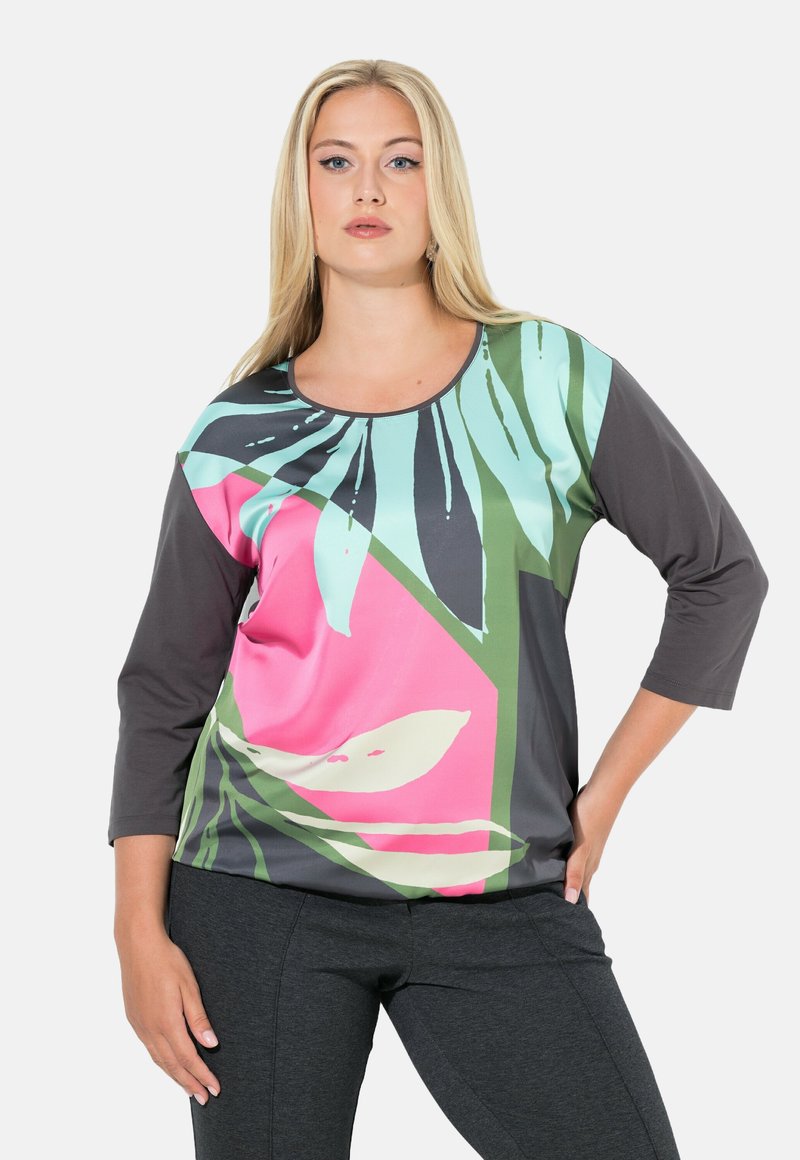 Vaaleatukkainen nainen, joka pukeutunut moniväriseen pitkähihaiseen yläosaan, jossa on vaaleanpunainen, vihreä ja sininen abstrakti lehtikuvio, sekä tummanharmaat housut.