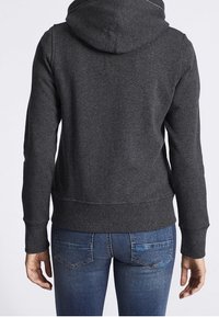 Sudadera gris con capucha, con dobladillo y puños acanalados, que presenta una textura suave. Se lleva con vaqueros azules de mezclilla, mostrando la vista trasera.