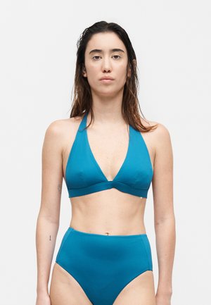 Γυναίκα με βρεγμένα μαλλιά που φοράει μια τιρκουάζ μπικίνι χαλτέρ με σλιπ ψηλόμεσο, στέκεται μπροστά σε λευκό, μονόχρωμο φόντο.