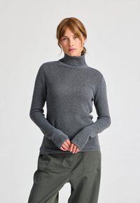 Pulover gri cu guler înalt, realizat dintr-un material ribbed, având mâneci lungi cu manșete extinse, purtat cu pantaloni cargo verzi.