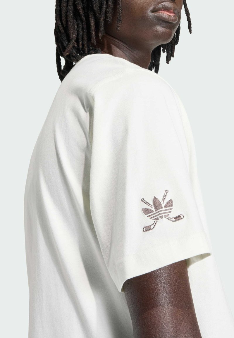 Vit bomullst-shirt med avslappnad passform. Har en brun broderad logotyp på vänster ärm. Mjuk textur och minimalistisk design.