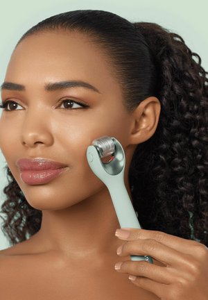 Femme aux cheveux bouclés utilisant un rouleau derma vert clair sur sa joue droite pour un traitement de soin de la peau.