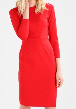 Kobieta w dopasowanej, czerwonej sukience do kolan z rękawami do łokci, stojąca na jednolitym białym tle.