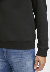 Sudadera de algodón negra con puños y dobladillo de canalé, combinada con jeans de mezclilla azules. Presenta una pequeña etiqueta de la marca en el lateral.