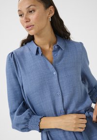 Chemise bleu clair à boutons avec des manches bouffantes, des poignets smockés et un tissu texturé. Présente un col et un détail de taille nouée.