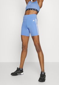 Personne athlétique portant un short bleu Adidas, un soutien-gorge de sport assorti, des chaussettes noires Adidas et des chaussures de sport noires, se tenant avec les bras levés.