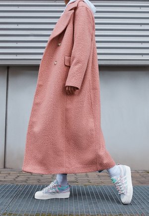 Dlouhý růžový texturovaný kabát s vystřiženým klopem a dvěma knoflíky. V kombinaci s vícebarevnými teniskami a bílými ponožkami.