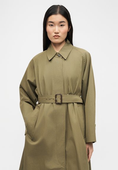 Ασιάτισσα με μακριά μαύρα μαλλιά, ντυμένη με χακί πράσινο παλτό με ζώνη, στέκεται με το ένα χέρι στην τσέπη μπροστά σε απλό φόντο.
