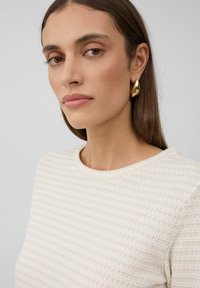 Pull blanc texturé avec un col rond, complété par de grandes boucles d'oreilles en or en forme de goutte. Fond neutre avec un éclairage doux.