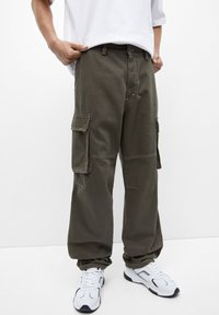 Person in olivgrünen Cargo-Hosen mit Seitentaschen und weißen Sneakern, der vor einem schlichten weißen Hintergrund steht.