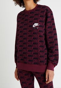 Bordeaux stickad sweatshirt med svart upprepad mönster av "NIKE" och "AIR"; har ribbade ärmslut och nederkant, rund halsringning och liten logotyp på bröstet.