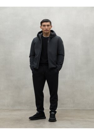 Chaqueta acolchada negra con capucha, llevada sobre un suéter negro, combinada con pantalones de chándal negros y zapatillas negras sobre un fondo neutro.
