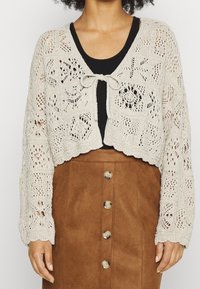 Cárdigan de ganchillo en beige claro con patrones de encaje floral, diseño corto y lazo en la parte delantera. Combinado con una falda corta de ante marrón con botones.