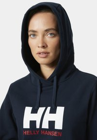 Námořnická mikina s kapucí, která má velké bílé logo "HH" a text "HELLY HANSEN" v červené barvě. Měkká látka s hladkým povrchem a nastavitelnými šňůrkami.