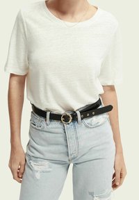 Donna che indossa una camicia bianca a maniche corte, jeans chiari strappati e una cintura nera con una fibbia circolare dorata.