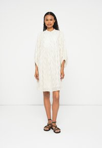 Vestido branco com padrão em zig-zag e cintura franzida, com mangas balão e decote redondo, usado com sandálias pretas de tiras.