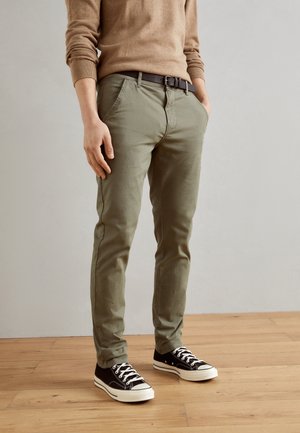 Person trägt olivgrüne Slim-Fit-Hose, schwarzen Gürtel, schwarz-weiße Sneaker und einen beigen Pullover und steht auf einem Holzboden.