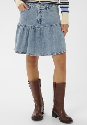 Femeie purtând o fustă de denim albastru, asortată cu cizme de piele maro până la jumătatea gambei, stând cu o mână în buzunar pe un fundal alb.