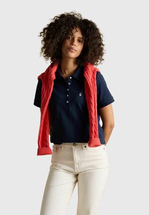 Frau mit lockigem Haar, die ein marineblaues Polohemd, weiße Hose und einen roten Strickpullover, der über ihre Schultern gelegt ist, vor einem schlichten Hintergrund trägt.