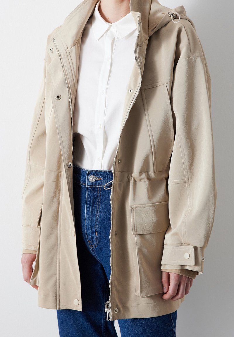 Jasnobeżowa kurtka na zamek z kapturem i dużymi kieszeniami. Wykonana z teksturowanej tkaniny, z zatrzaskami i luźnym krojem.