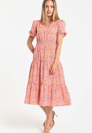 Woman standing in a pink midi dress with puff sleeves and a leopard print, wearing beige high-heeled sandals against a white background.