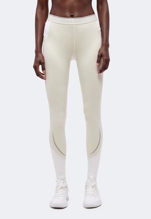 Leggings beige clair taille slim avec poches latérales, portés avec des chaussures de sport blanches sur une personne debout sur un fond uni.