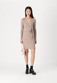 Robe portefeuille en tricot beige à côtes, à manches longues, décolleté en V et ceinture à nouer, assortie à des chaussures à plateforme noires et un petit sac à main blanc.