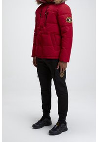 Doudoune rouge avec une capuche en fourrure, poches zippées et un patch logo ; associée à un pantalon noir avec poches cargo et détails.