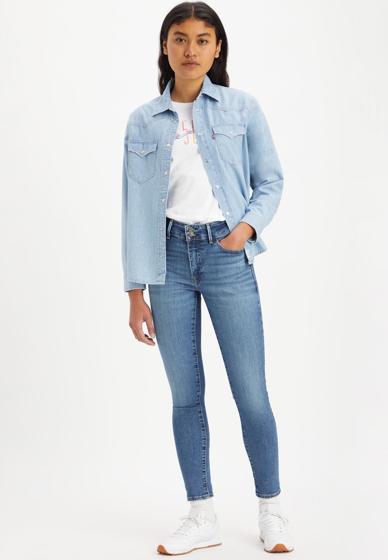 Chemise en denim bleu clair avec deux poches avant, associée à un jean bleu ajusté. Baskets blanches et t-shirt graphique en dessous.