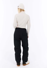 Zwarte broek met een relaxte pasvorm en elastische tailleband, gecombineerd met een licht beige langemouwen shirt en een crèmekleurige gebreide beanie, gezien van achteren.