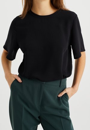 Femme portant un chemisier noir à manches courtes rentré dans un pantalon taille haute vert foncé, les mains dans les poches.