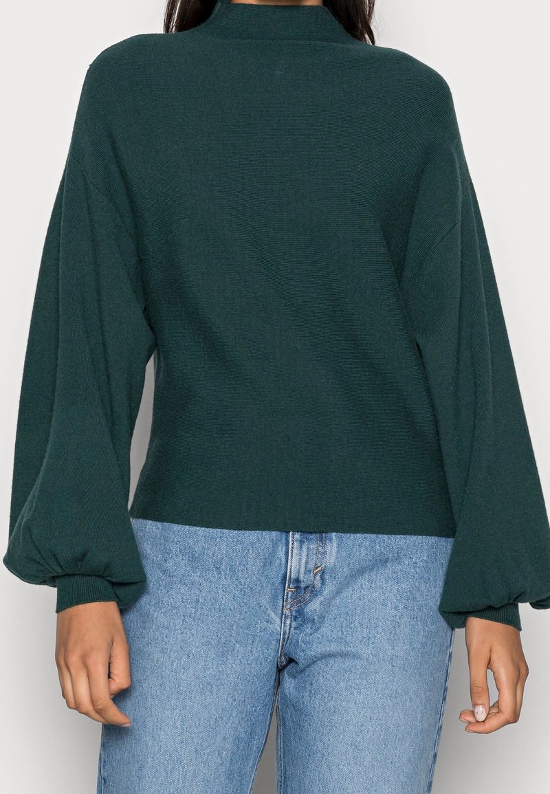 Femme portant un pull à manches longues vert foncé avec des poignets bouffants et un jean taille haute bleu clair, debout devant un fond uni.