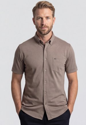 Mann mit hellbraunen Haaren und Bart, der ein kurzärmeliges taupefarbenes Hemd mit Brusttasche trägt und mit den Händen in den Taschen steht.