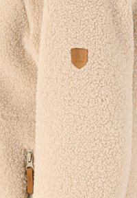 Beige fleecejakke med en fyldig tekstur, der har en lynlås og et brunt læderlogo. Blødt, hyggeligt stof med et varmt udseende.