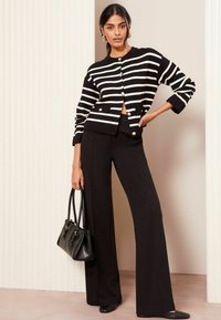 Zwart-wit gestreepte cardigan met gouden knopen, gecombineerd met hoge zwarte broek. Het model draagt een zwarte handtas.