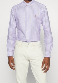 Chemise boutonnée rayée violet clair avec des lignes blanches et lavande, ornée d'un petit logo brodé, associée à un jean blanc.