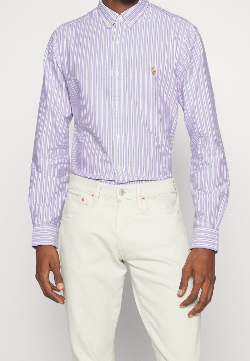 Chemise boutonnée rayée violet clair avec des lignes blanches et lavande, ornée d'un petit logo brodé, associée à un jean blanc.