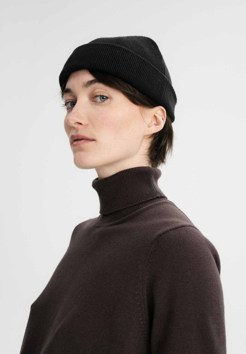 Zwarte geribbelde beanie op een model met kort donker haar, gecombineerd met een aansluitende bruine coltrui. Duidelijke focus op texturen en kleuren.
