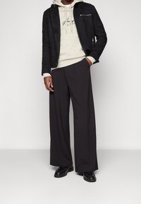 Chaqueta de ante negra, sudadera crema con logo, pantalones amplios negros y zapatos negros. Enfoque en la textura y colores contrastantes.