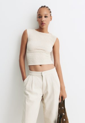 Beige textured crop top with a round neckline, paired with cream pleated trousers. Model holds a brown shoulder bag with metallic accents.