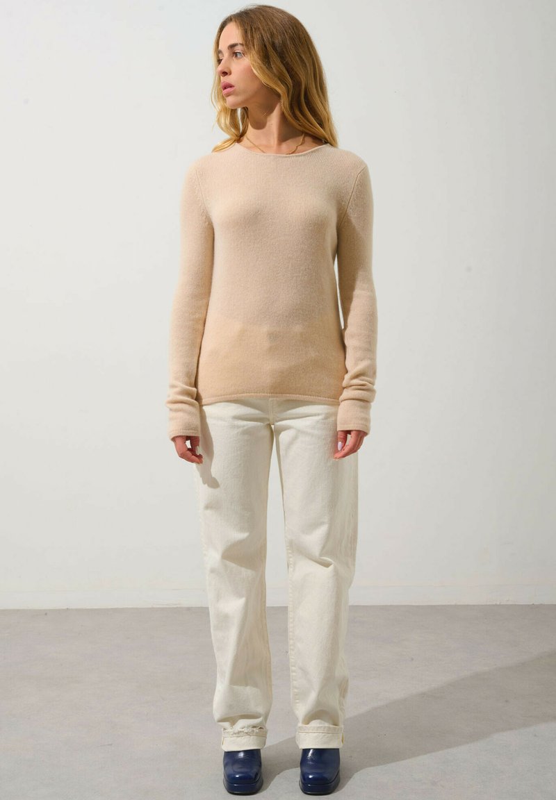 Beige Strickpullover mit langen Ärmeln und gerippten Kanten, kombiniert mit weißen weit geschnittenen Jeans und navy-farbenen Plateauschuhen. Glatte Textur.
