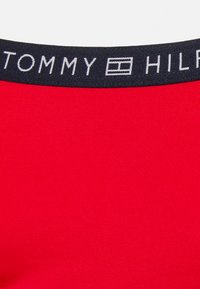 Tecido vermelho em um corte justo, com uma faixa elástica preta apresentando o nome da marca "TOMMY HILFIGER" em texto branco.