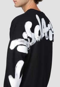 Vista laterale di una persona che indossa una felpa nera con testo e disegni in stile graffiti, in bold bianco, sulla schiena e sulla manica.